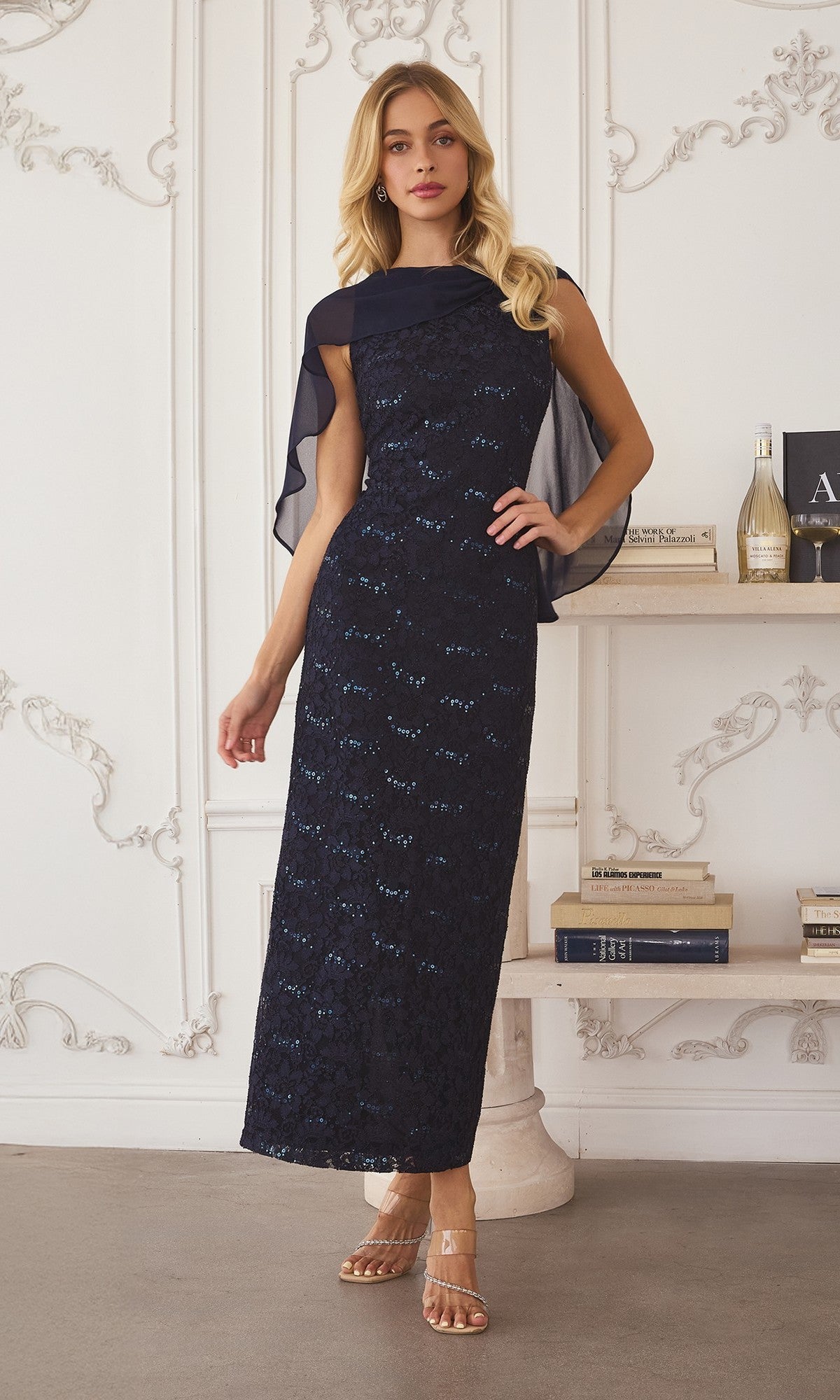 Woman in the Long Prom Dress: Amoris T906—dark blue sequin-lace with sheer shoulder detail—stands indoors by a bookshelf and champagne.